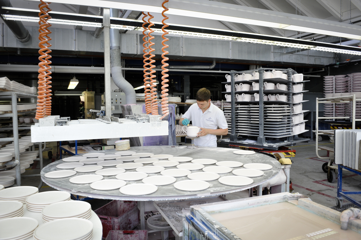 Mitarbeiter glasiert Porzellanteller in Fabrikhalle, Arbeitsfläche voller Teller. Fürstenberg Porzellan.