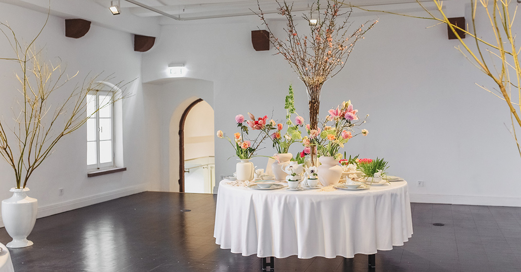 Rund gedeckter Tisch mit Blumenarrangement, in hell erleuchtetem Raum. Fürstenberg Porzellan.