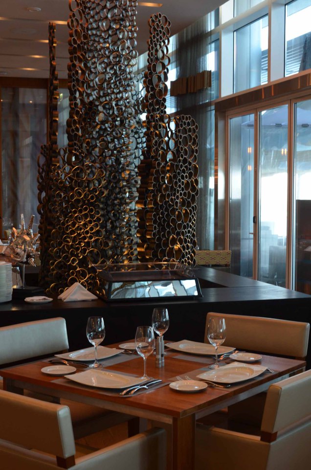 Gedeckter Holztisch mit Gläsern, vor kunstvoller Skulptur aus Metallrohren im Restaurant. Fürstenberg Porzellan.