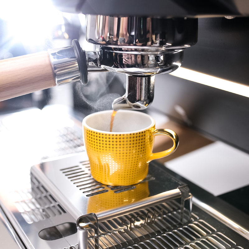 Gelbe Tasse unter Espressomaschine, Kaffee fließt ein, dampfend auf Metallplatte. Fürstenberg Porzellan.