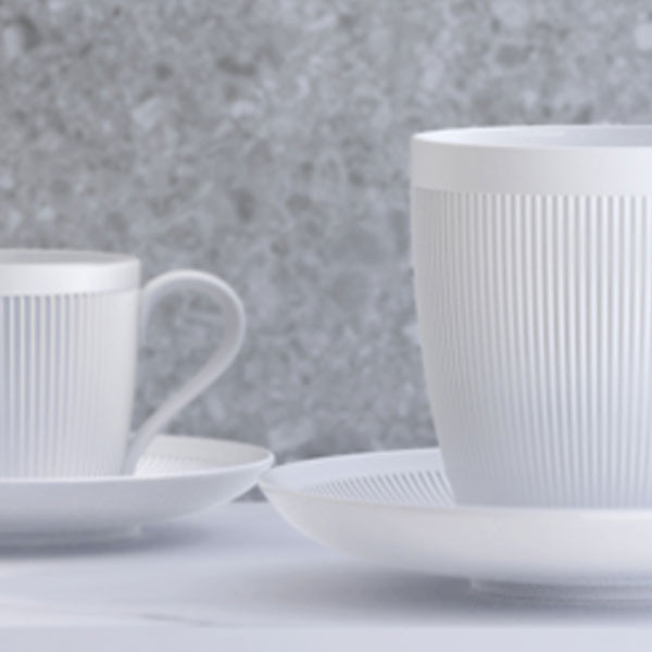 Weiße Porzellantassen mit vertikalen Rillen stehen auf Untertassen vor grauer Wand. Fürstenberg Porzellan.