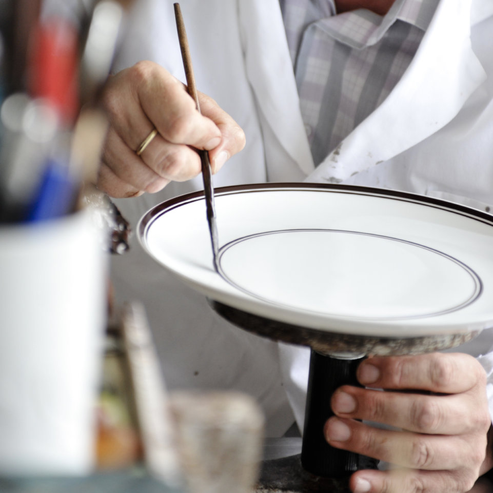 Hand bemalt Porzellanteller mit Pinsel, Person trägt weißen Kittel. Fürstenberg Porzellan.