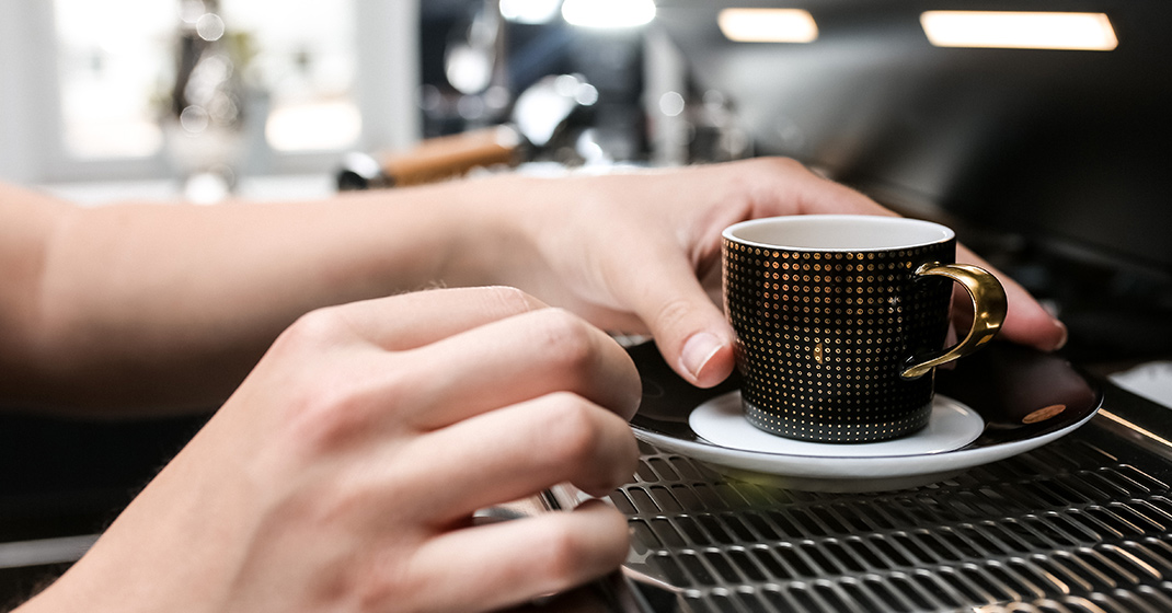 Hände positionieren schwarze Espressotasse mit Goldgriff auf Untertasse neben Espressomaschine. Fürstenberg Porzellan.