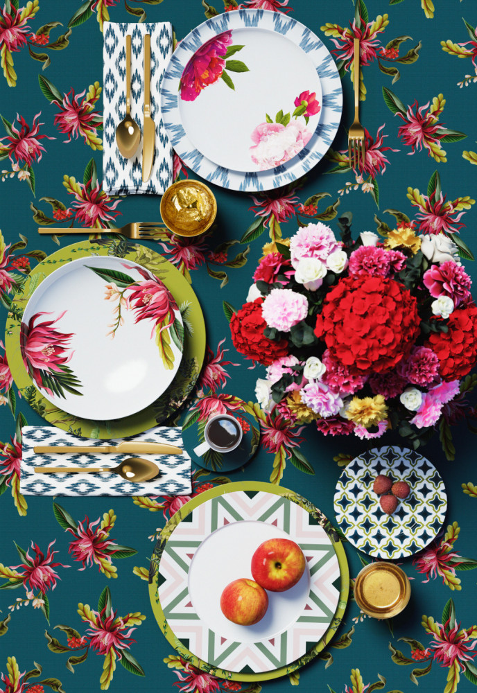 Bunte Teller mit Blumen- und Obstmotiven arrangieren sich auf floralem Tischdeckenhintergrund. Fürstenberg Porzellan.