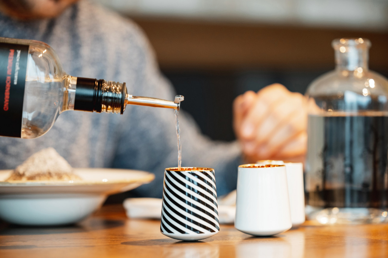 Flasche gießt Flüssigkeit in gestreifte Tasse auf Tisch neben weißer Tasse. Fürstenberg Porzellan.