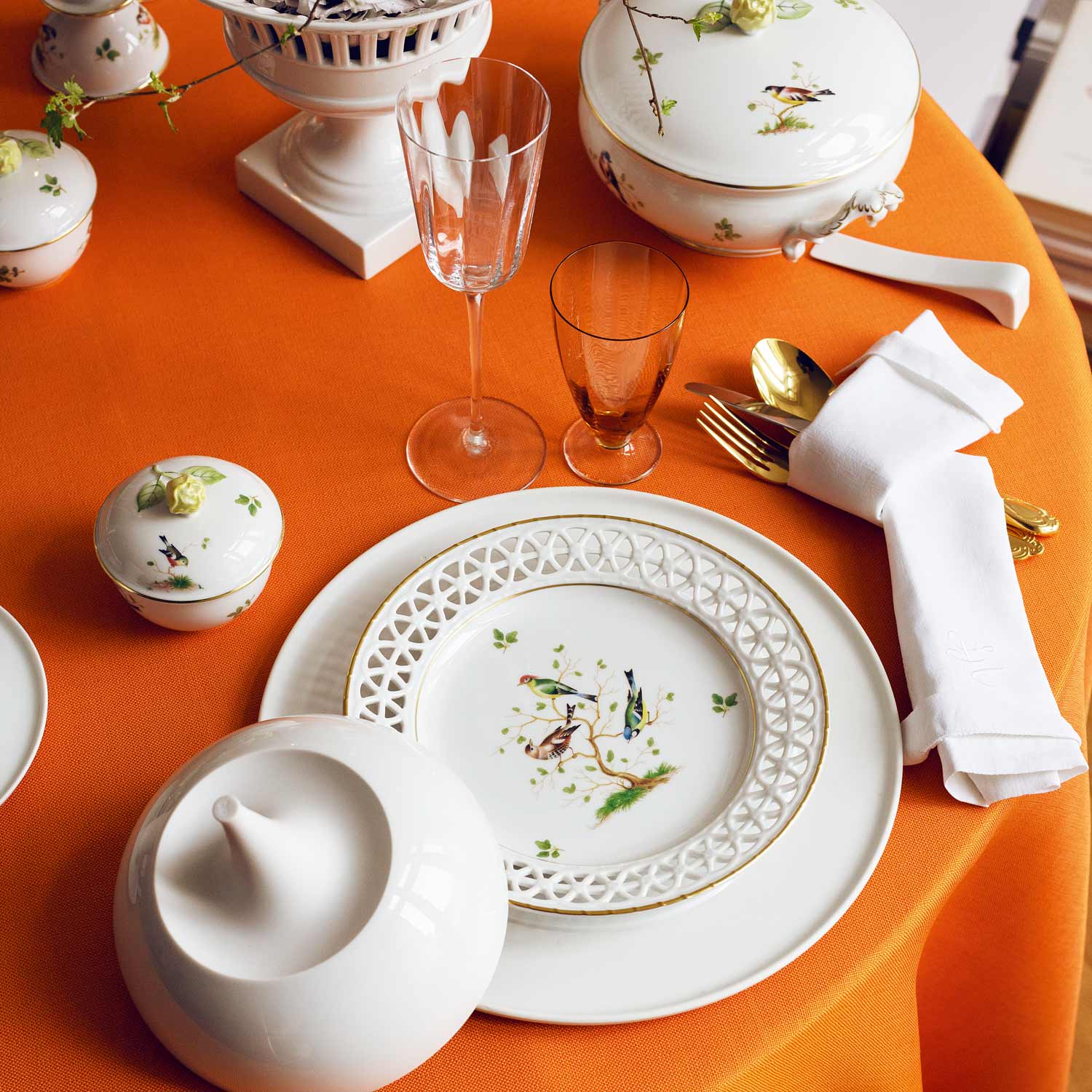 Porzellanteller mit Vogelmotiv und Blumendekor auf orangefarbener Tischdecke arrangiert. Fürstenberg Porzellan.
