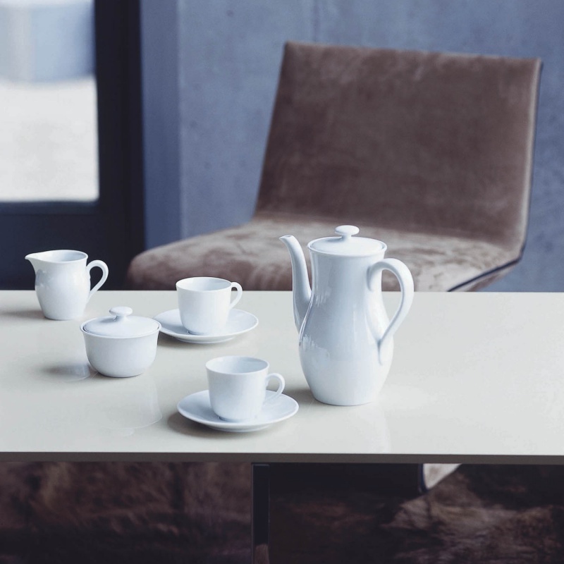 img2_ta_6395_weiss_kaffeetasse_2_tlg Weiße Porzellan-Kaffeeservice-Garnitur auf einem Tisch mit modernem Stuhl im Hintergrund. Fürstenberg Porzellan.