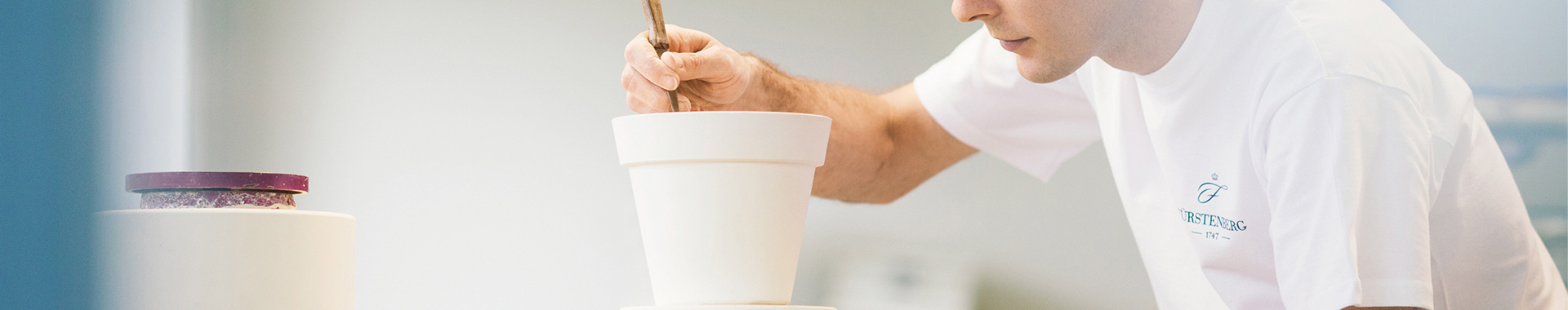 INDUSTRIEKERAMIKER (M/W/D) · FACHRICHTUNG VERFAHRENSTECHNIK Person bemalt Porzellantopf mit Pinsel, konzentriert im hellen Arbeitsraum. Fürstenberg Porzellan.