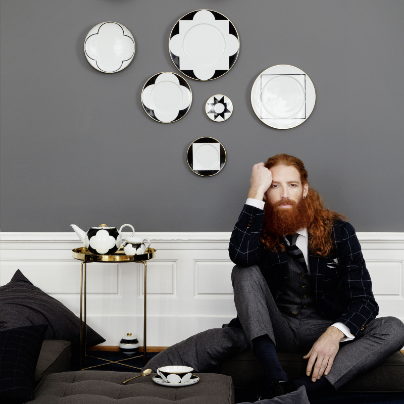 img5_fl2006235705_fruehstuecksteller Mann sitzt auf Sofa, daneben Teeservice auf Tisch, Porzellanteller an Wand. Fürstenberg Porzellan.