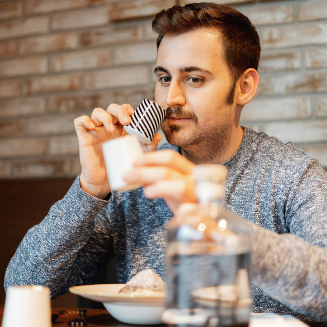 Mann riecht Streifentasse, hält weiße Tasse, sitzt vor Backsteinwand, Essen vor ihm. Fürstenberg Porzellan.