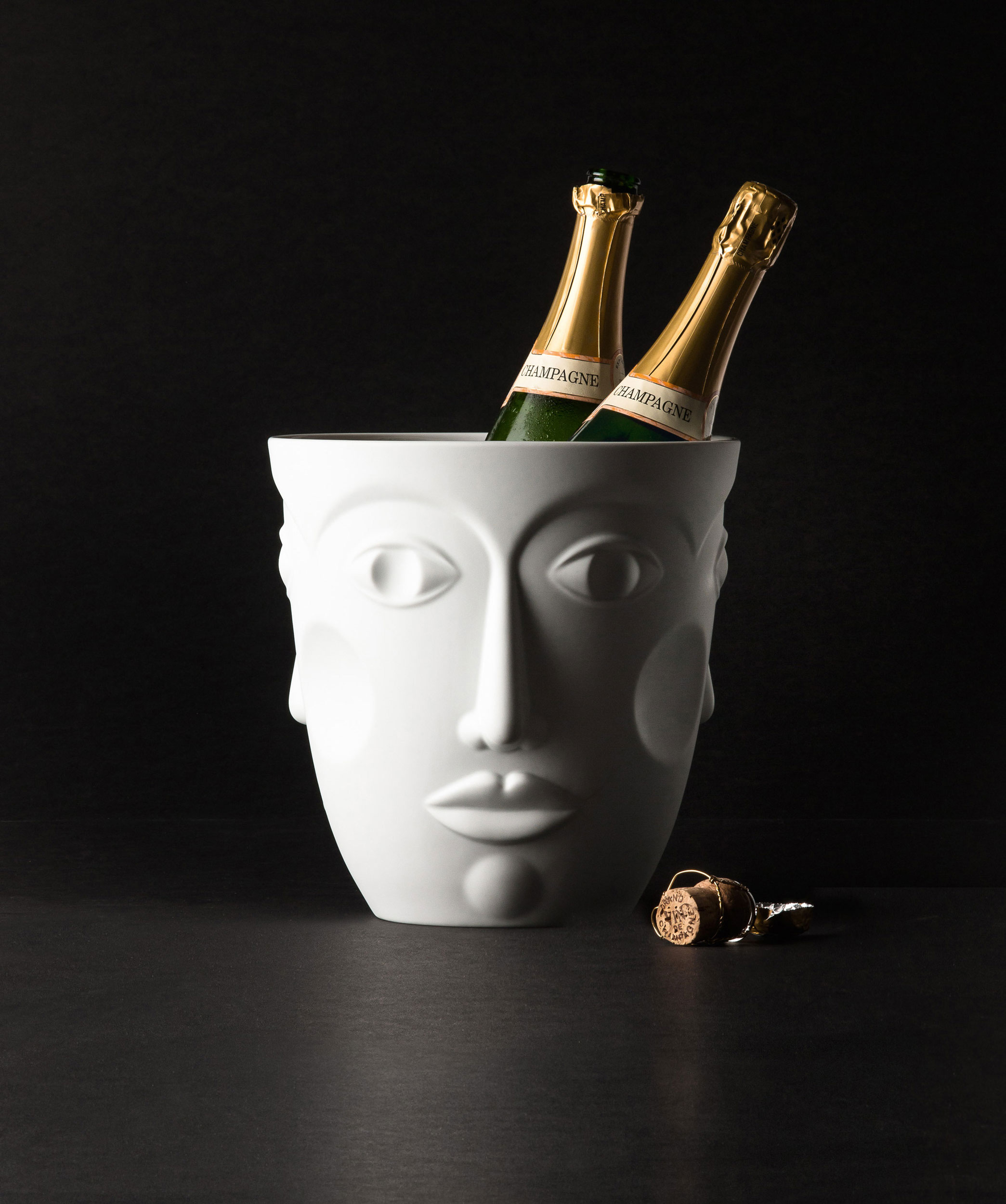 Weißer Gesichtskübel hält zwei Champagnerflaschen, daneben liegt ein Sektkorken. Fürstenberg Porzellan.