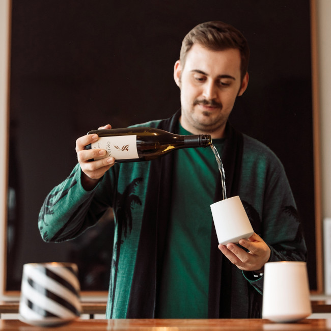 Mann gießt Wein in weißes Gefäß, steht vor dunklem Hintergrund. Fürstenberg Porzellan.