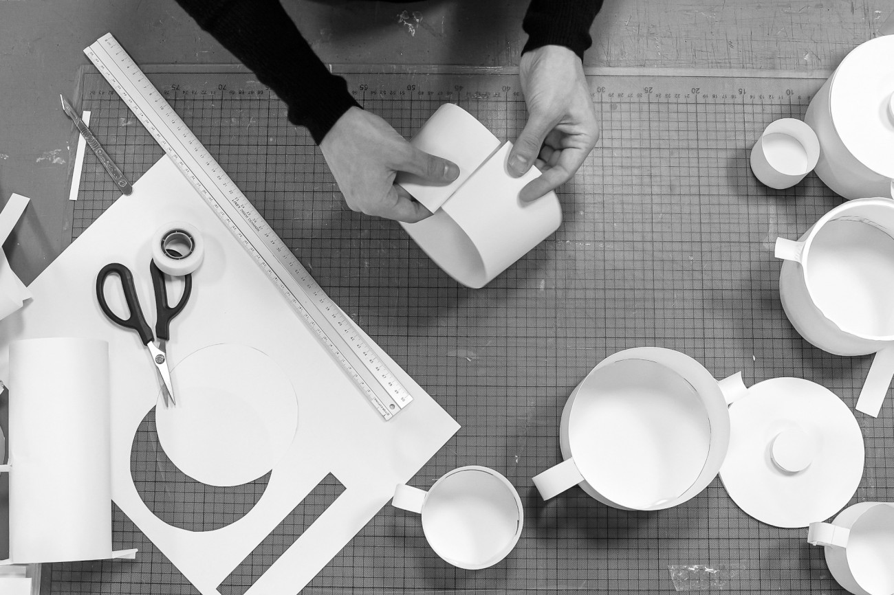 Hände falten Papiermodell von Tasse auf Schreibtisch mit Lineal und Schere. Fürstenberg Porzellan.