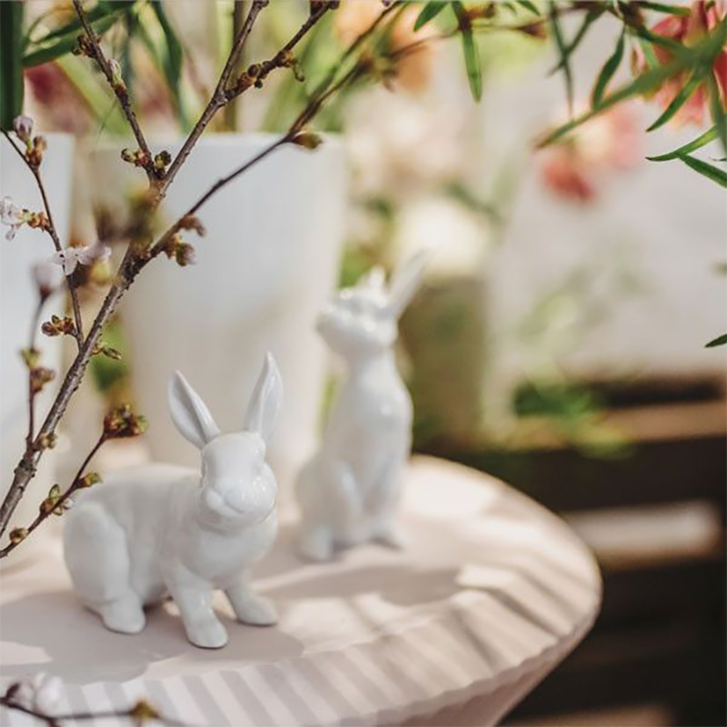 Zwei weiße Hasenfiguren sitzen auf Tisch neben Zweigen und Blumen. Fürstenberg Porzellan.