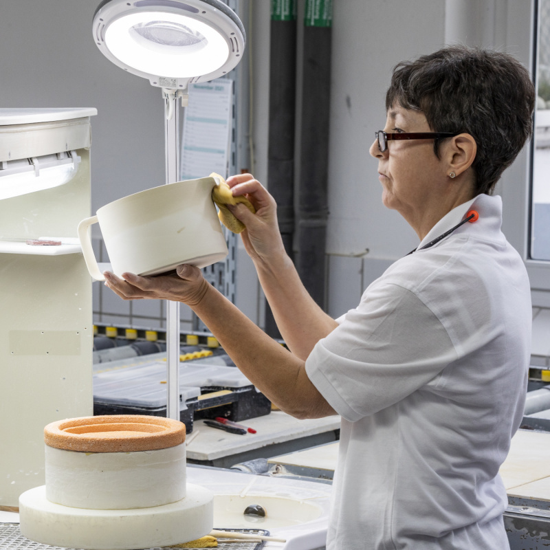 img3_tk20183_1001_teekanne Person reinigt Porzellanobjekt mit Schwamm unter Lampe in Werkstatt. Fürstenberg Porzellan.