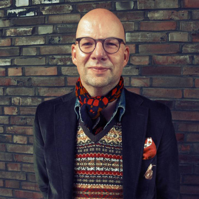 Mann trägt Brille, gemusterten Pullover, Schal, steht vor Ziegelwand. Fürstenberg Porzellan.