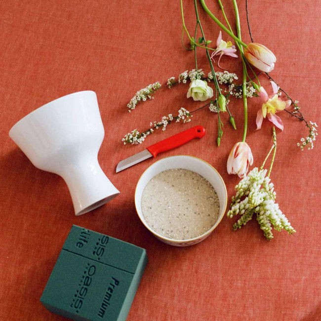 Weiße Vase, Schale mit Sand und Blumen liegen auf rotem Tuch. Fürstenberg Porzellan.