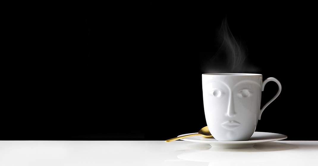 Weiße Kaffeetasse mit Gesichtsmotiv dampft, steht auf Untertasse mit goldenem Löffel. Fürstenberg Porzellan.