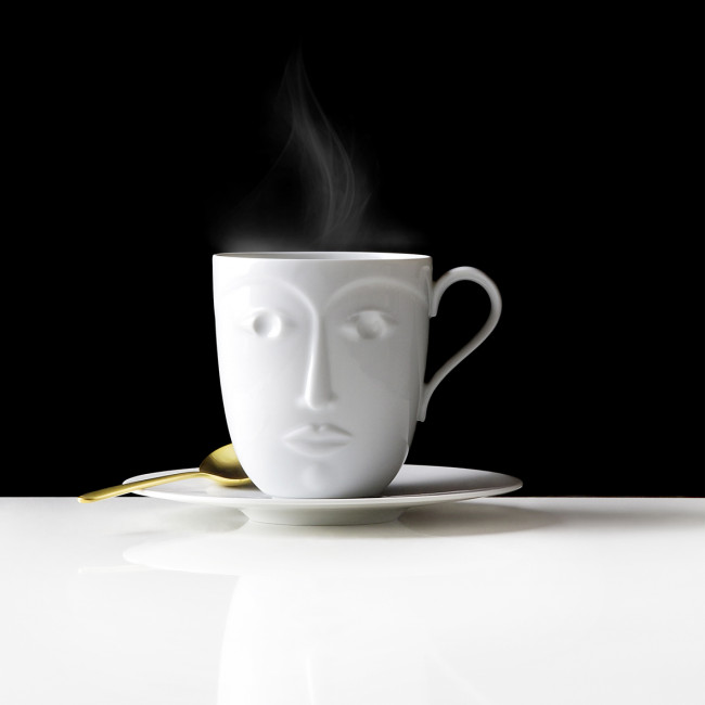 Weiße Tasse mit Gesichtsmotiv, dampfend, auf Untertasse mit goldfarbenem Löffel. Fürstenberg Porzellan.