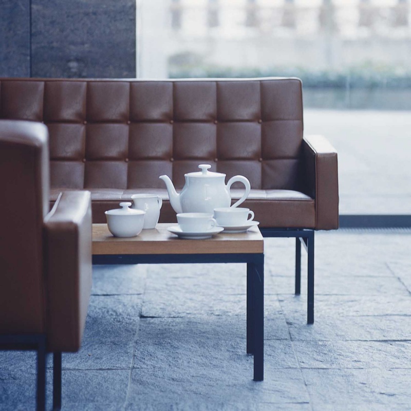 img2_un_6395_weiss_kaffee__teeuntertasse Weiße Teekanne mit Tassen auf Holztisch, braune Ledersofas im Hintergrund. Fürstenberg Porzellan.