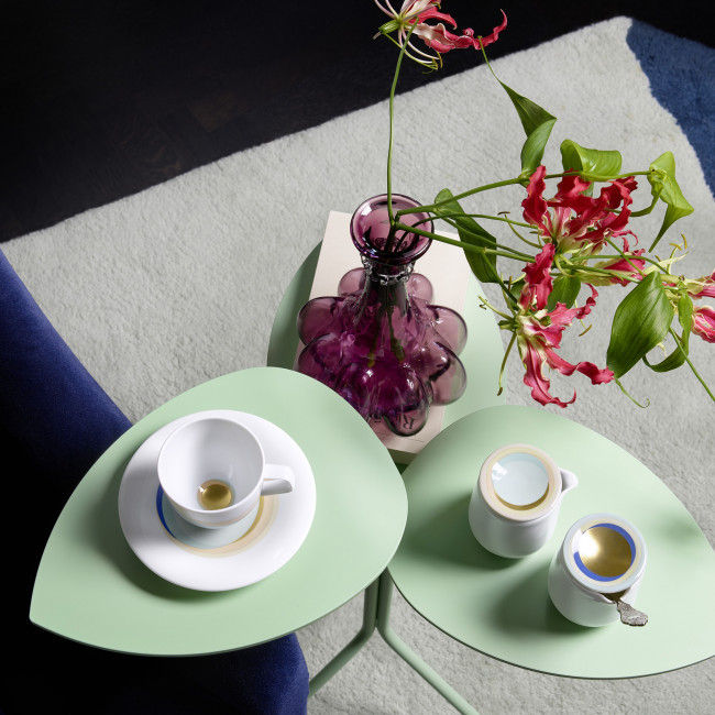 Weißes Teeservice und lila Blumenvase auf grünem Tisch vor blauem Sofa. Fürstenberg Porzellan.