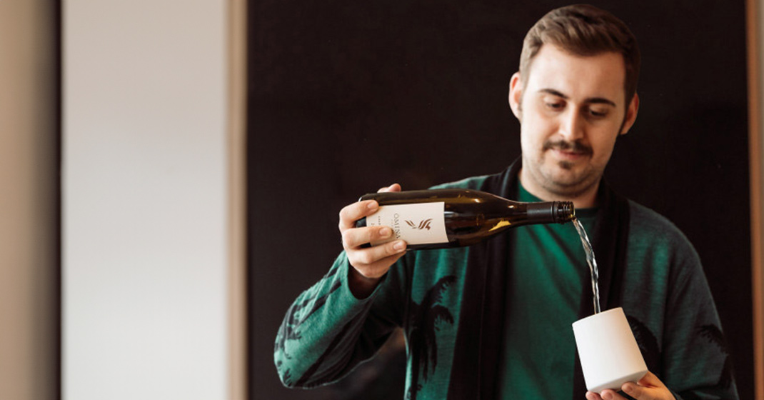 Mann gießt aus Flasche in Becher, lächelnd vor dunklem Hintergrund. Fürstenberg Porzellan.