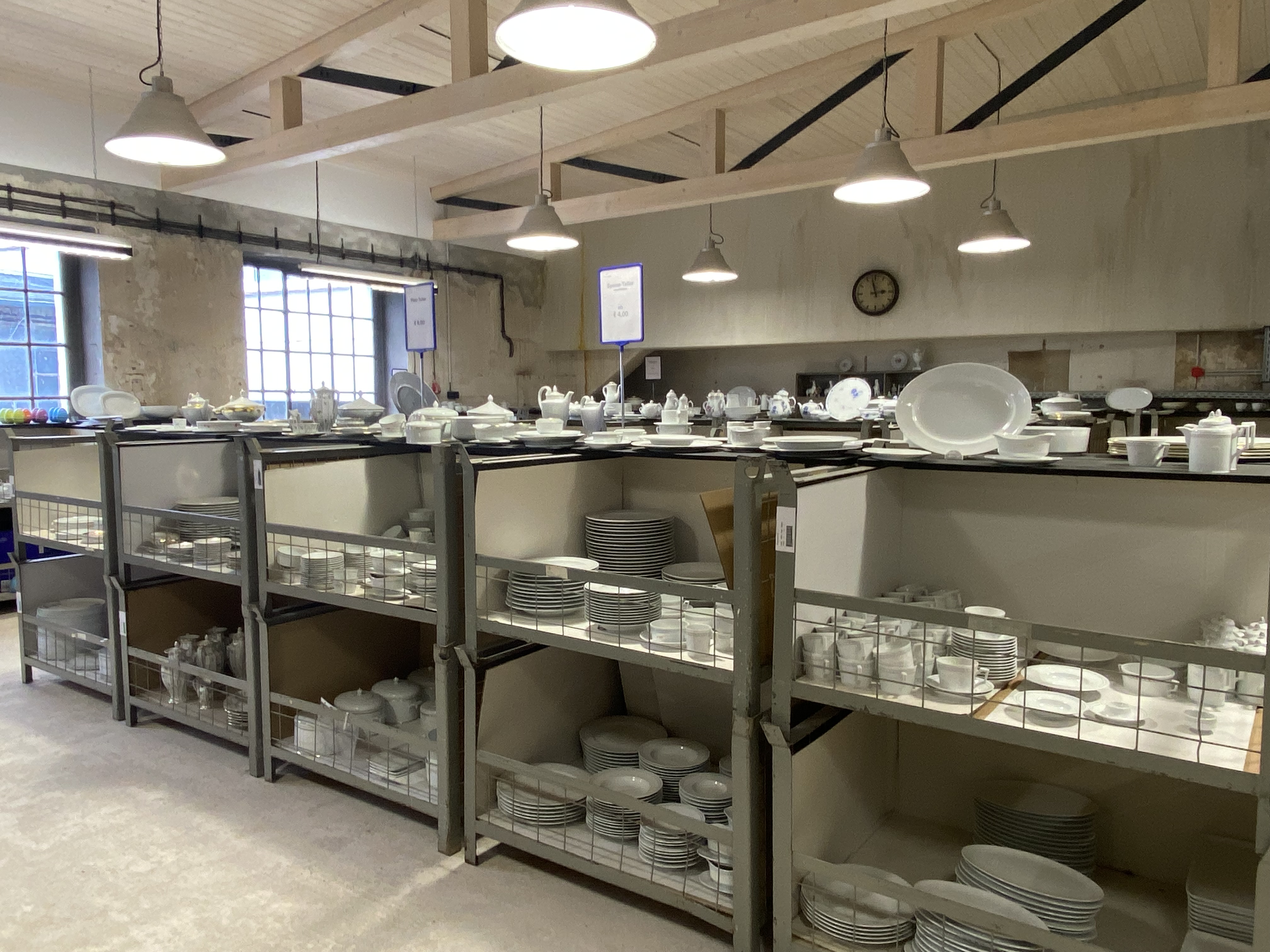 Porcelain special sale Regale mit weißem Porzellangeschirr in einem Lagerraum, beleuchtet von Hängelampen. Fürstenberg Porzellan.