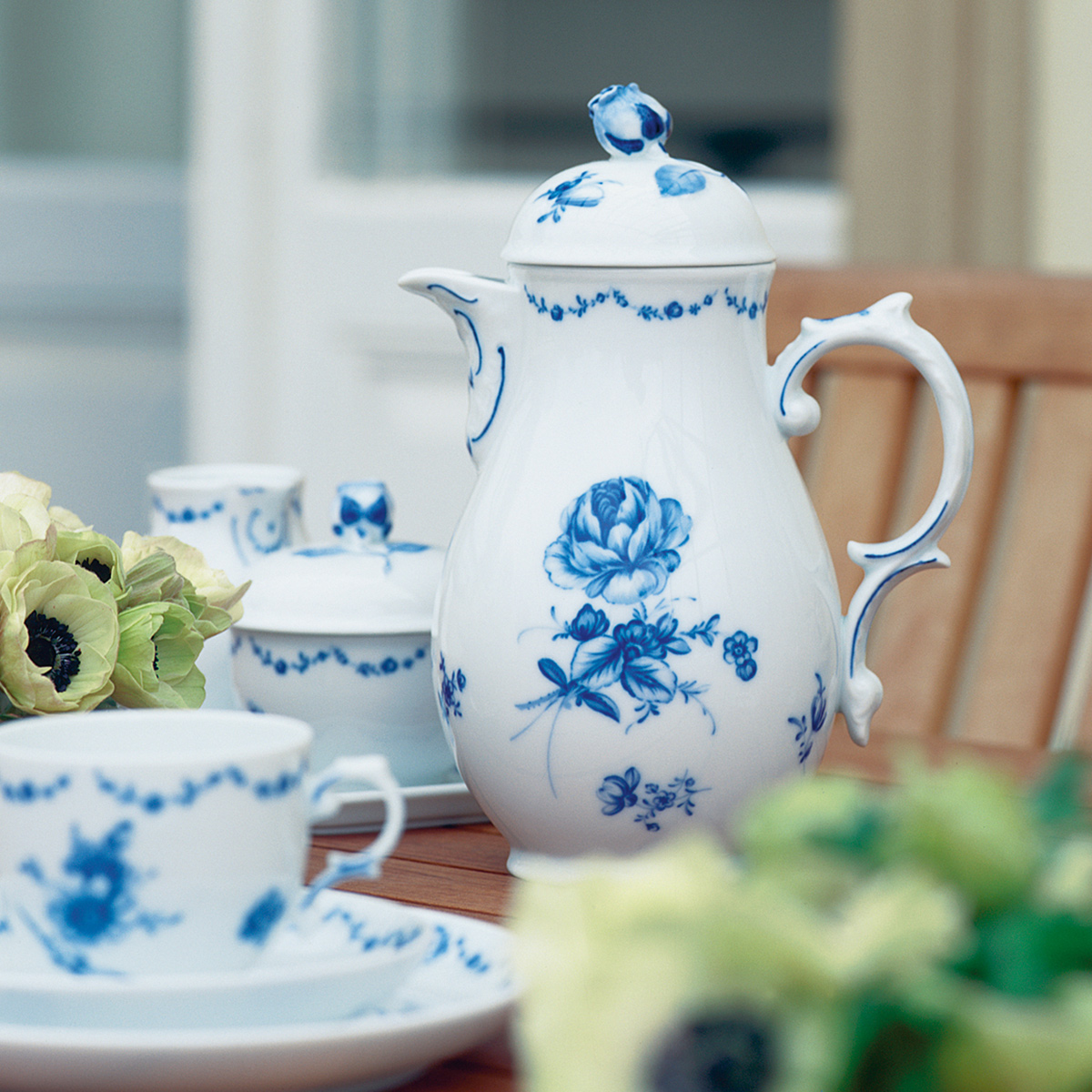Porzellankanne mit blauen Blumenmotiven steht auf Tisch, umgeben von Geschirr und Blumen. Fürstenberg Porzellan.