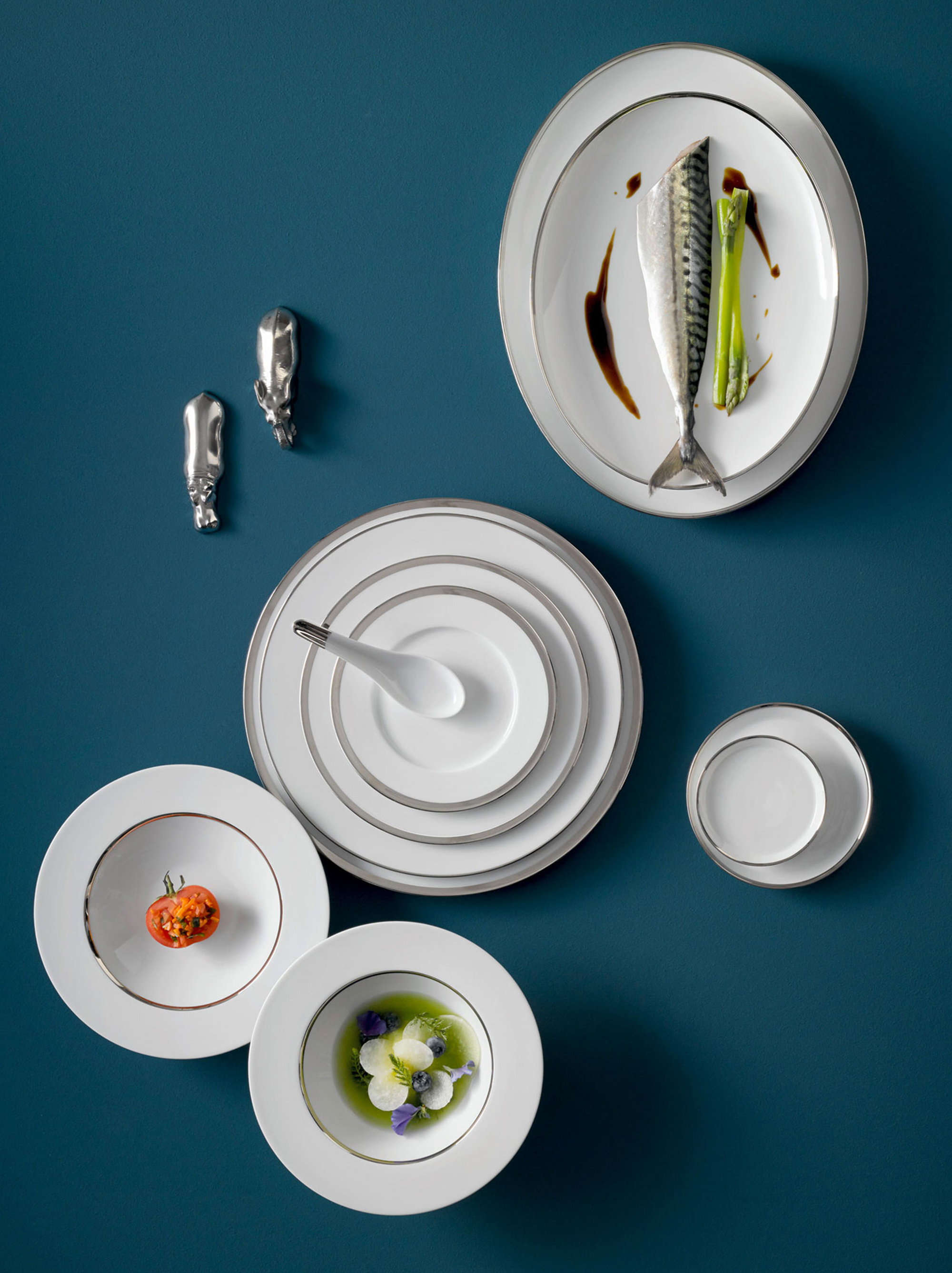 Weißes Porzellan-Geschirr auf blauem Hintergrund, mit Makrele, Spargel, Tomate arrangiert. Fürstenberg Porzellan.