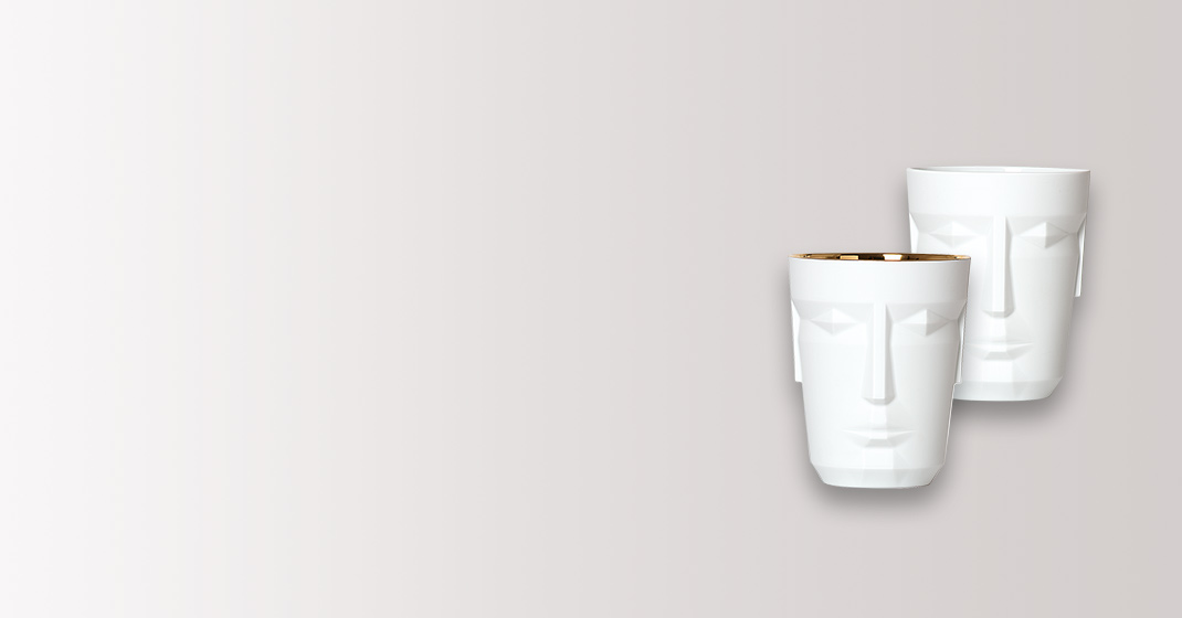 Zwei weiße Becher mit Gesichtsrelief, einer mit goldenem Rand, stehen nebeneinander. Fürstenberg Porzellan.