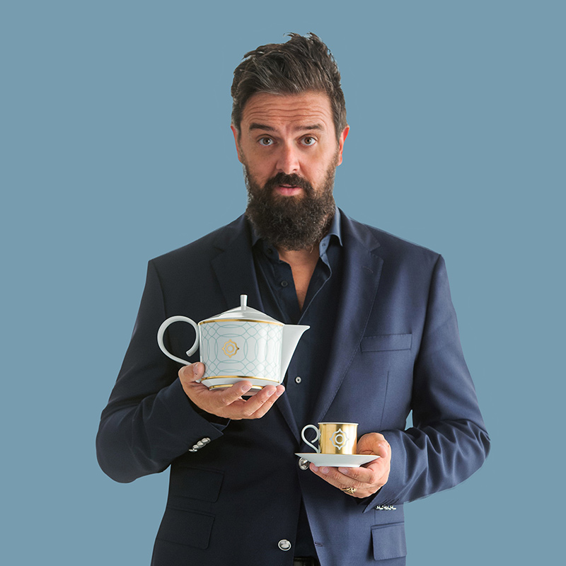 Mann in Anzug hält Porzellan-Teekanne und Tasse vor blauem Hintergrund. Fürstenberg Porzellan.