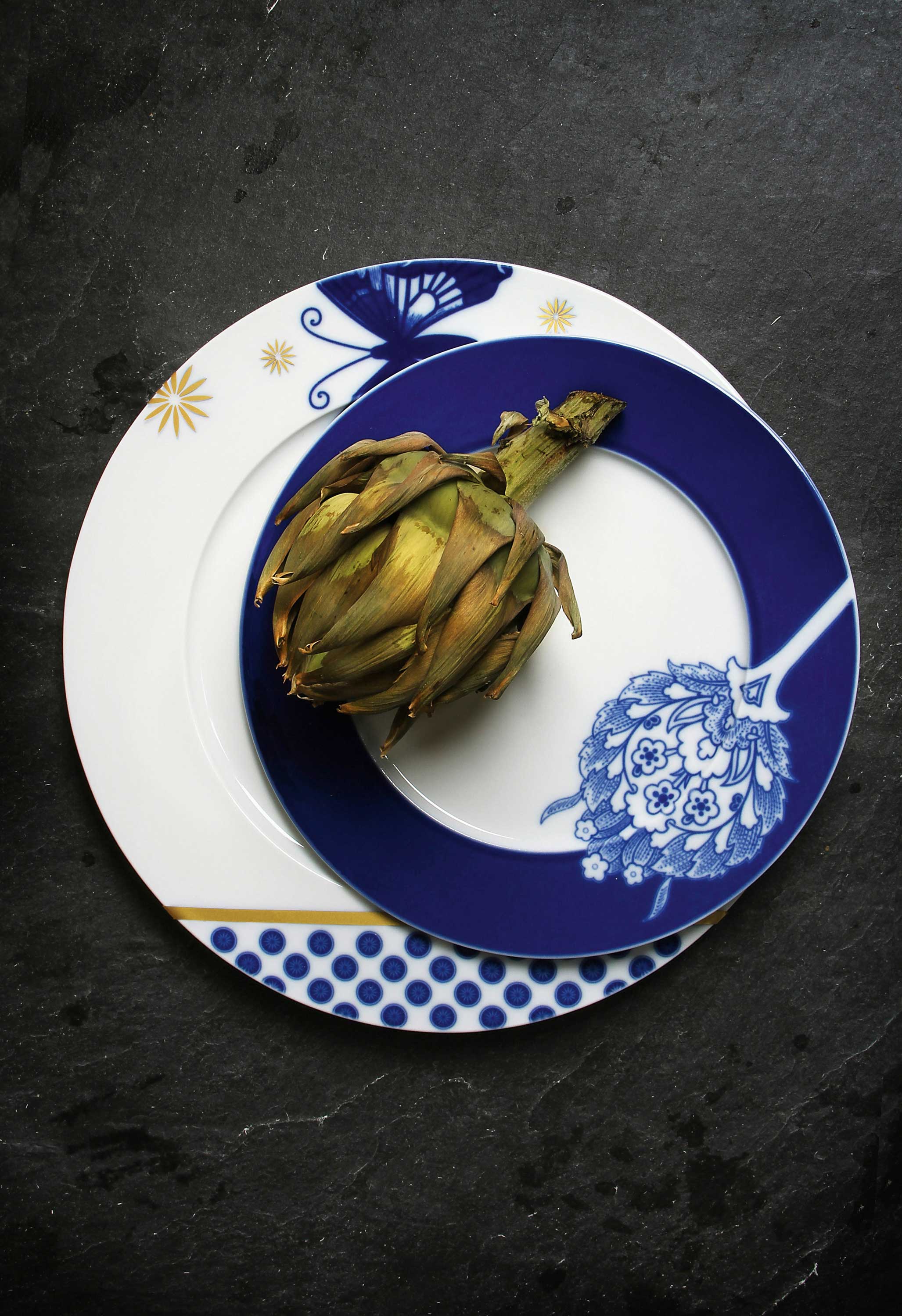 Artischocke liegt auf blau-weiß verziertem Teller mit Blumenmuster auf dunklem Hintergrund. Fürstenberg Porzellan.