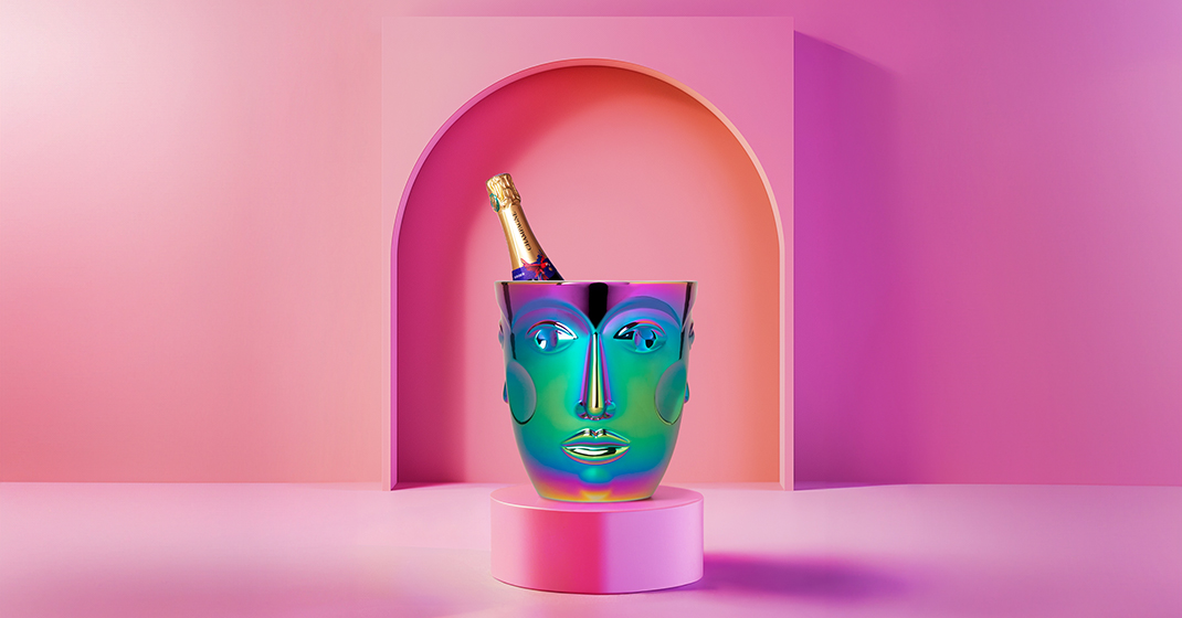 Champagnerflasche steckt in einem regenbogenfarbenen Porzellankühler mit Gesichtsmotiv, pinker Hintergrund. Fürstenberg Porzellan.