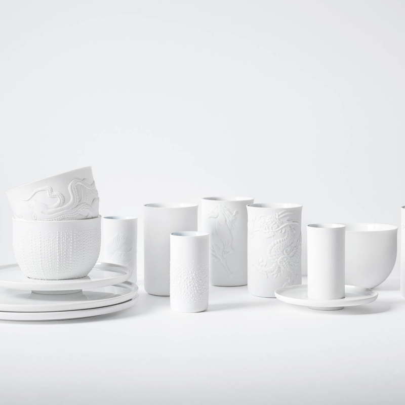 img3_obp8791_1002_becher_doppelwandig__relief_kugelfische_ Weißes Porzellan-Geschirr mit eingeprägten Mustern, auf gestapelten Tellern arrangiert. Fürstenberg Porzellan.